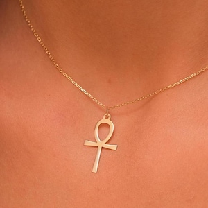 May include: A gold-colored Ankh pendant necklace. The Ankh symbol, a cross with a loop at the top, hangs from a delicate chain. The necklace is worn around the neck, with the pendant resting near the collarbone.