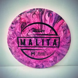 May include: A pink and purple disc golf disc with the word "MALITA" in bold black letters. The disc has a marbled appearance and the text "DISCRAFT" and "FIRST RUN" are also visible.
