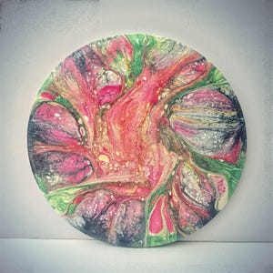 May include: Round abstract art piece with a fluid design. The artwork features a blend of colors, including pink, red, green, and black, creating a unique and eye-catching pattern. The piece is circular and appears to be made of a solid material.