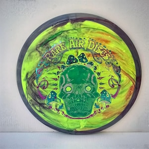 May include: A round, neon green disc with a dark gray rim. The disc features a green skull design with mushrooms and the text "RARE AIR DISCS" in a psychedelic font. The disc has a swirling, colorful pattern.