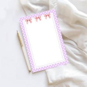 May include: A blank stationery sheet with a light purple checkered border and pink bow accents. A white pen with a gold-colored tip rests beside the stationery on a white surface. The stationery is designed for writing notes.