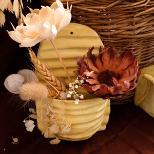 Könnte beinhalten: Dekorative Anordnung mit einem hellgelben Keramikbehälter mit geripptem Design, der getrocknete Blumen und Weizen enthält. Eine große, cremefarbene Blume und eine dunkelrote Blume sind prominent. Ein Weidenkorb befindet sich im Hintergrund.