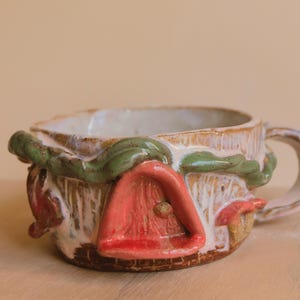 Può includere: Una tazza in ceramica stravagante con un design a casetta delle fate. La tazza presenta una porta rossa, tralci verdi e un interno color crema. Il manico è curvo e la tazza ha un aspetto rustico e fatto a mano.