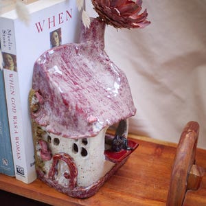 Può includere: Un vaso decorativo in ceramica a forma di casa, con tetto rosso e corpo bianco sporco strutturato. Un grande fiore bordeaux secco è nell'apertura del vaso. Il vaso è su una superficie di legno, vicino a libri con la parola "WHEN" visibile.