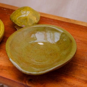 May include: An olive-green ceramic bowl with an organic shape and speckled texture. The bowl has a glossy finish. A small, rounded ceramic piece is behind the bowl. The items are on a wooden surface.