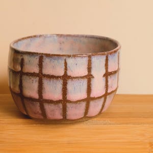 May include: A handcrafted ceramic bowl with a grid design. The bowl displays a gradient of pink and blue tones, with brown grid lines. The bowl is placed on a wooden surface, highlighting its unique design and craftsmanship.