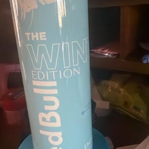 Puede incluir: Una lata de Red Bull azul claro con el texto "THE WIN EDITION" en blanco. La lata est&aacute; sobre un posavasos azul a juego. El logotipo de Red Bull se muestra prominentemente en blanco. La lata tiene un dise&ntilde;o con tem&aacute;tica invernal.