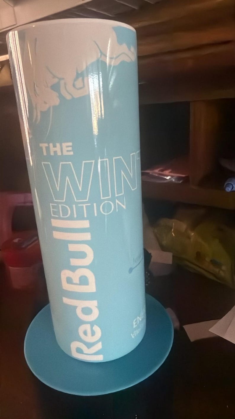 Puede incluir: Una lata de Red Bull azul claro con el texto "THE WIN EDITION" en blanco. La lata est&aacute; sobre un posavasos azul a juego. El logotipo de Red Bull se muestra prominentemente en blanco. La lata tiene un dise&ntilde;o con tem&aacute;tica invernal.