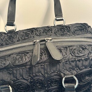May include: Black handbag with a rose pattern. The bag has a double zip closure and two handles with silver-coloured metal rings. The bag's exterior is covered in a textured, floral design.