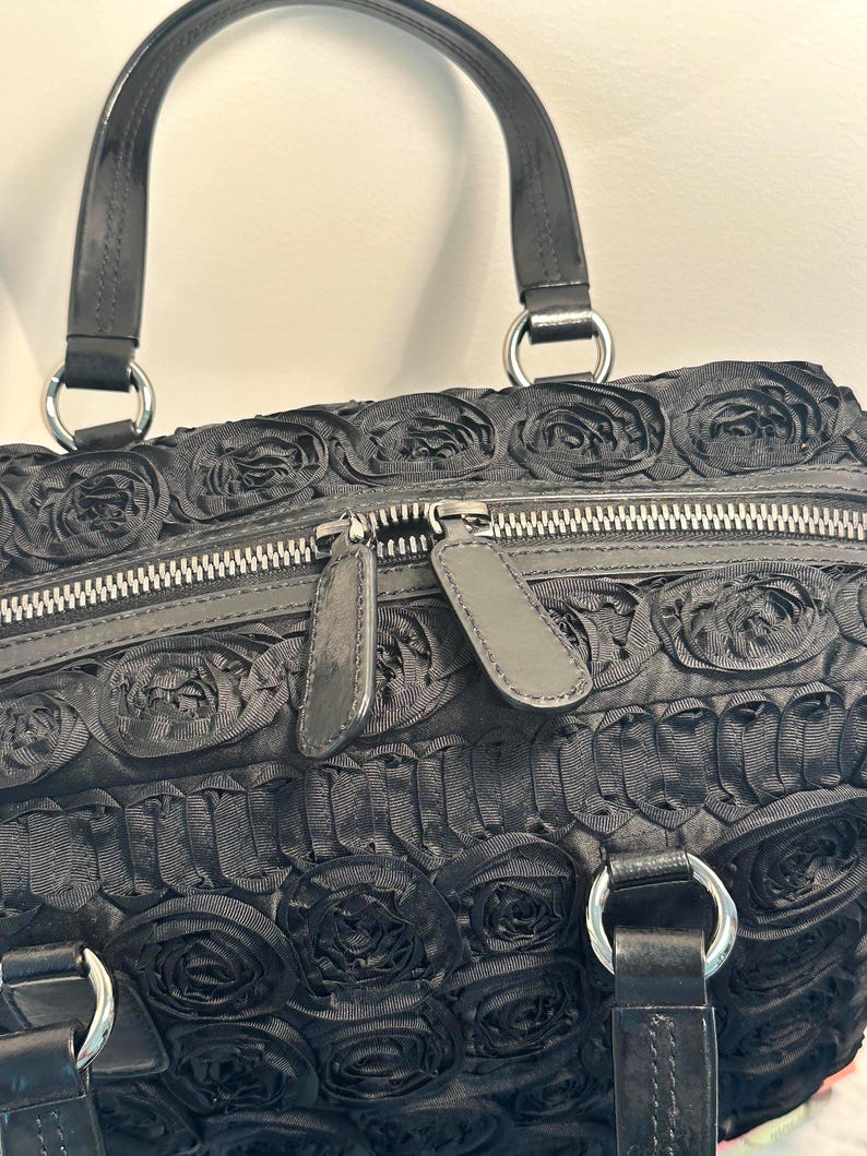 May include: Black handbag with a rose pattern. The bag has a double zip closure and two handles with silver-coloured metal rings. The bag's exterior is covered in a textured, floral design.