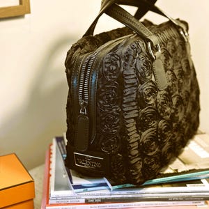 May include: Black handbag with a rose design, featuring a top handle and a zip closure. The bag has a silver Valentino logo and is resting on a stack of magazines.