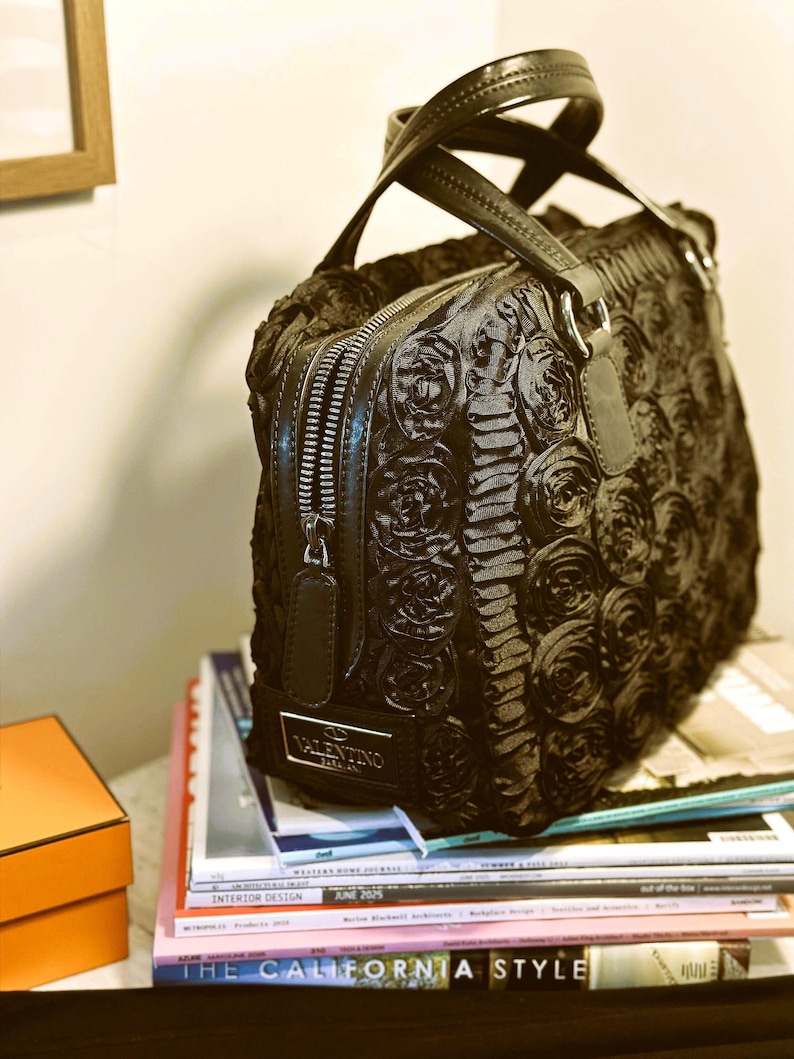 May include: Black handbag with a rose design, featuring a top handle and a zip closure. The bag has a silver Valentino logo and is resting on a stack of magazines.