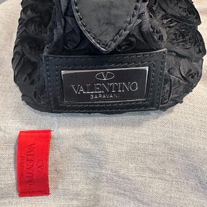 May include: Black Valentino Garavani handbag with a floral pattern. The bag has a silver logo plate and a black zip. A red Valentino tag is visible.
