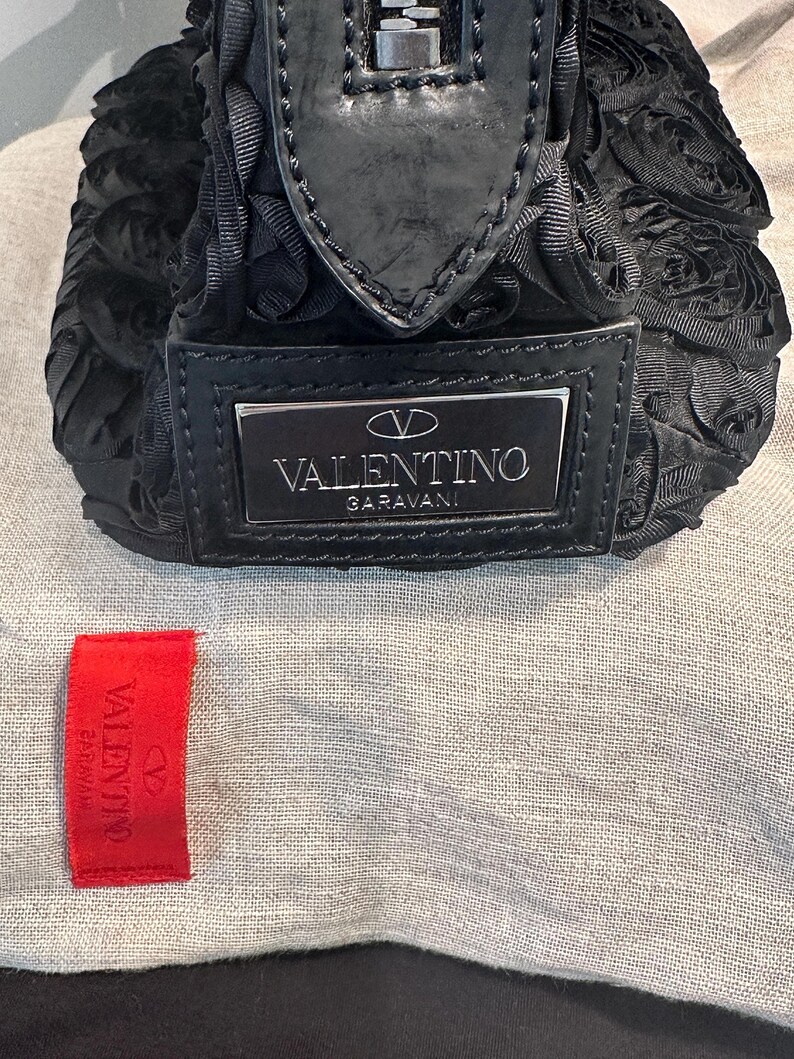 May include: Black Valentino Garavani handbag with a floral pattern. The bag has a silver logo plate and a black zip. A red Valentino tag is visible.