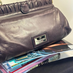 May include: A dark purple leather handbag with a top handle and gold-colored hardware. The bag features a rectangular metal plaque with the "Valentino Garavani" logo. The bag is resting on a stack of magazines.