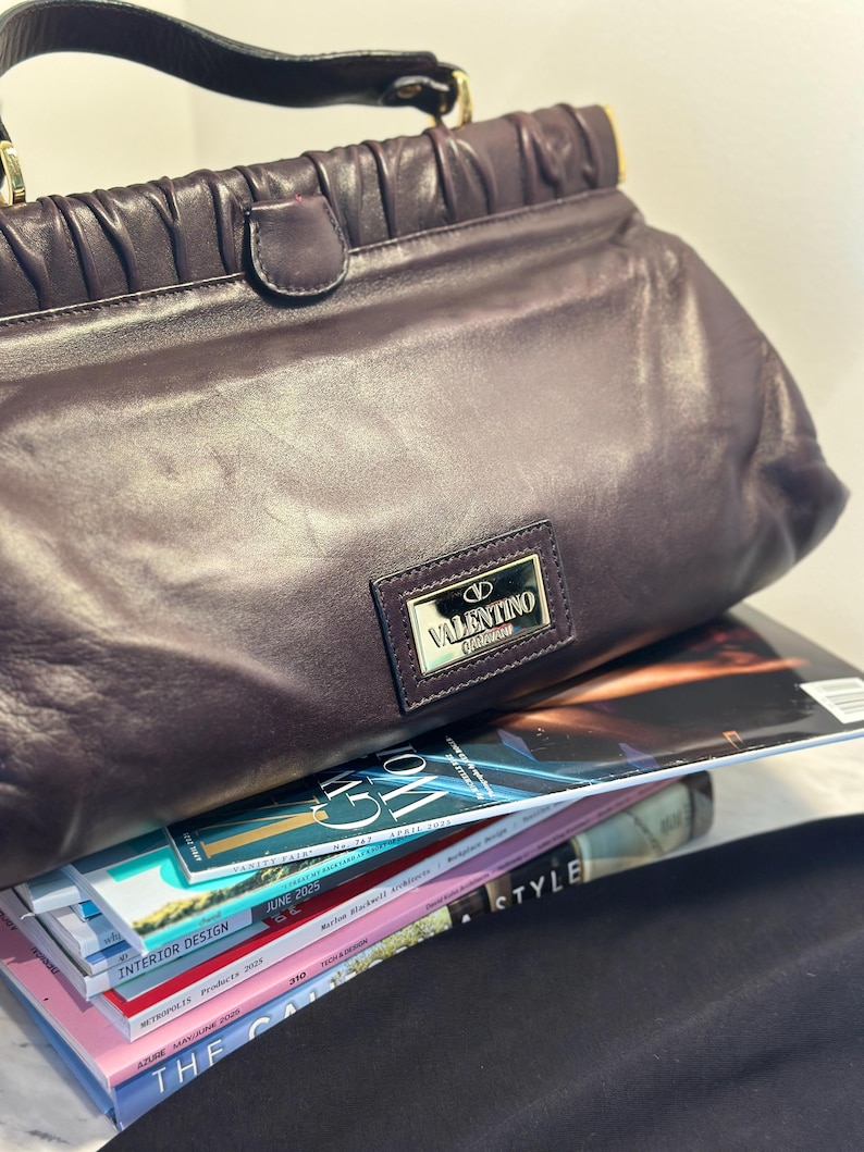 May include: A dark purple leather handbag with a top handle and gold-colored hardware. The bag features a rectangular metal plaque with the "Valentino Garavani" logo. The bag is resting on a stack of magazines.
