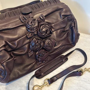 May include: A dark brown leather handbag with a decorative rose design. The bag has a shoulder strap with gold-colored clasps. The bag's surface is textured and pleated, with a top handle.