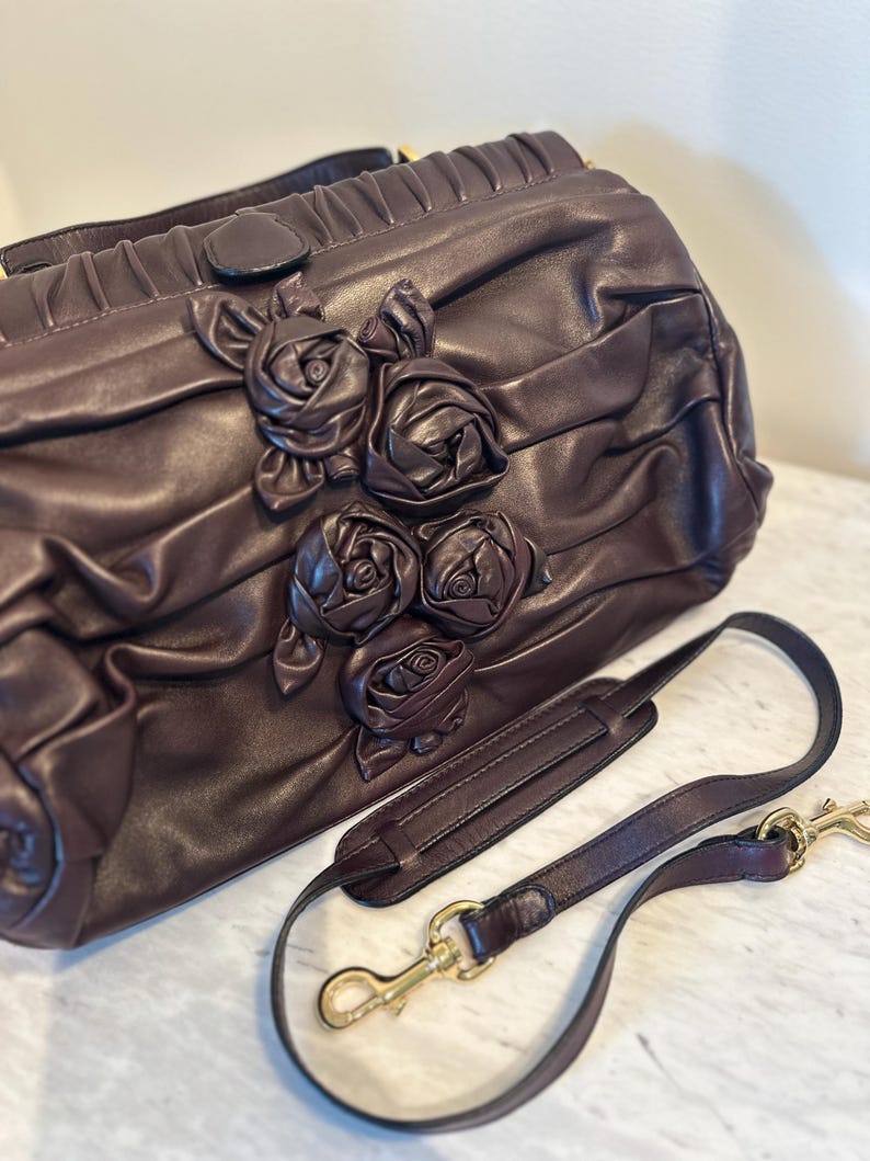 May include: A dark brown leather handbag with a decorative rose design. The bag has a shoulder strap with gold-colored clasps. The bag's surface is textured and pleated, with a top handle.