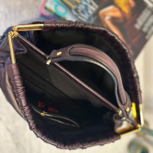 May include: A deep purple handbag with gold-colored metal accents. The bag has a drawstring closure and a top handle. The interior is black, with a small zippered pocket. A magazine is visible in the background.