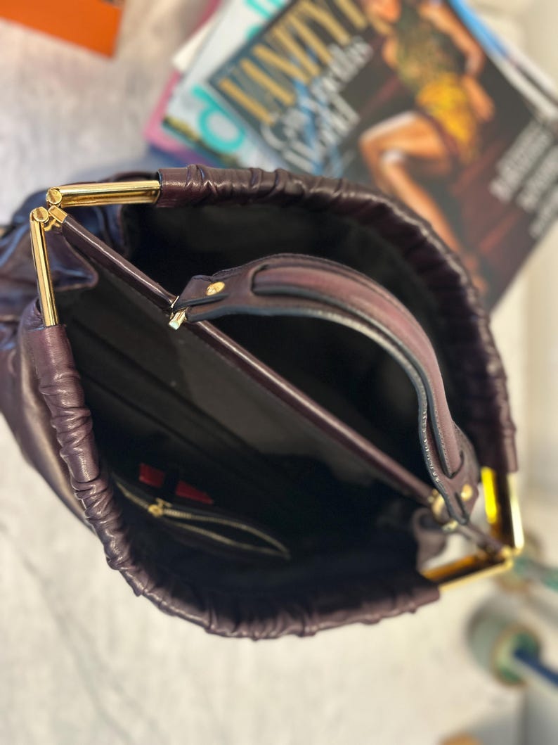 May include: A deep purple handbag with gold-colored metal accents. The bag has a drawstring closure and a top handle. The interior is black, with a small zippered pocket. A magazine is visible in the background.
