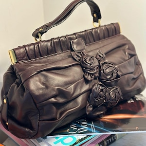 May include: A dark brown leather handbag with a top handle and decorative rose embellishments. The bag features a ruched design and gold-toned hardware. The handbag is sitting on top of a stack of magazines.