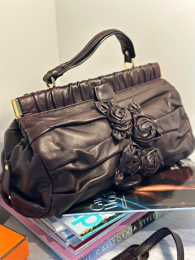 May include: A dark brown leather handbag with a top handle and decorative rose embellishments. The bag features a ruched design and gold-toned hardware. The handbag is sitting on top of a stack of magazines.