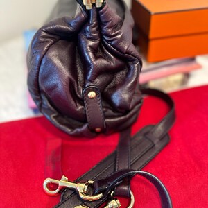 May include: A dark burgundy leather handbag with gold-tone hardware. The bag features a gathered design and a detachable shoulder strap with gold clasps. The bag is on a red surface.