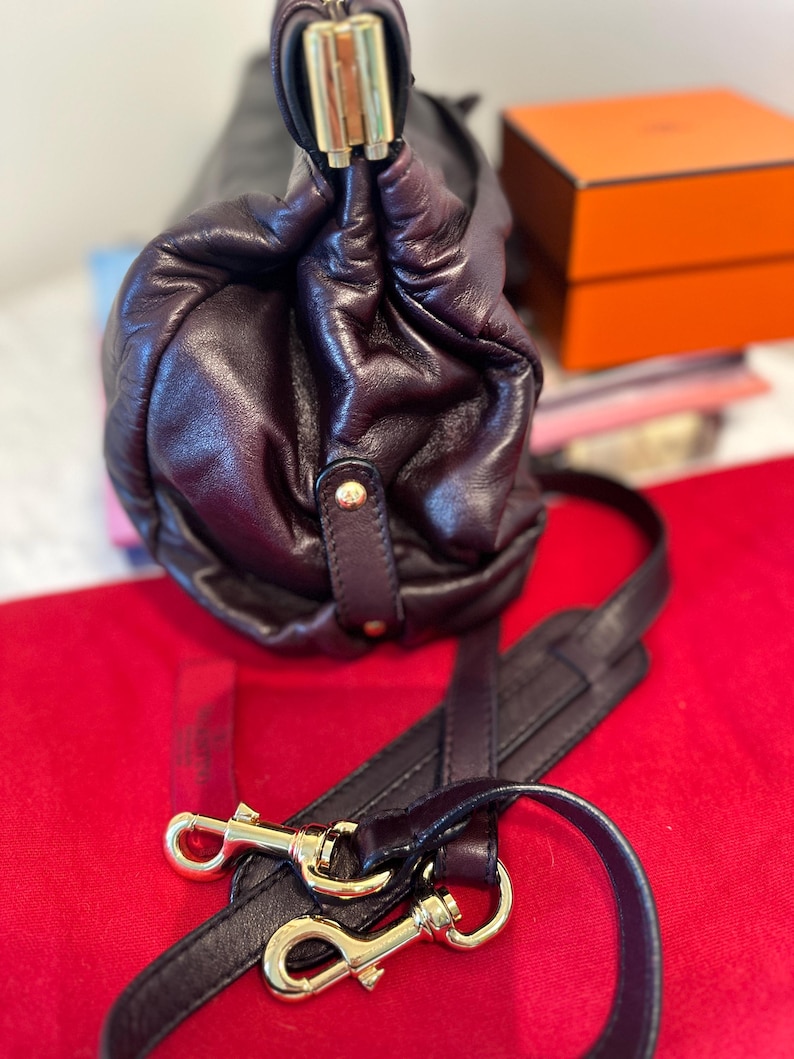 May include: A dark burgundy leather handbag with gold-tone hardware. The bag features a gathered design and a detachable shoulder strap with gold clasps. The bag is on a red surface.