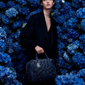 May include: A person in a black outfit holds a navy blue handbag with a rose pattern. The bag has two handles and silver hardware. The background is a wall of vibrant blue hydrangea flowers.