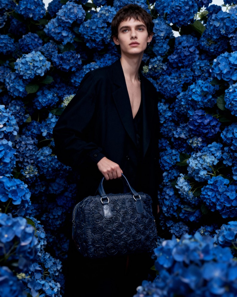 May include: A person in a black outfit holds a navy blue handbag with a rose pattern. The bag has two handles and silver hardware. The background is a wall of vibrant blue hydrangea flowers.