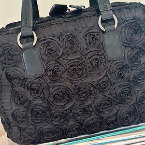 May include: Black handbag featuring a rose pattern. The bag has two handles with silver rings and a zip closure. It rests on a stack of magazines. The magazines have various titles.
