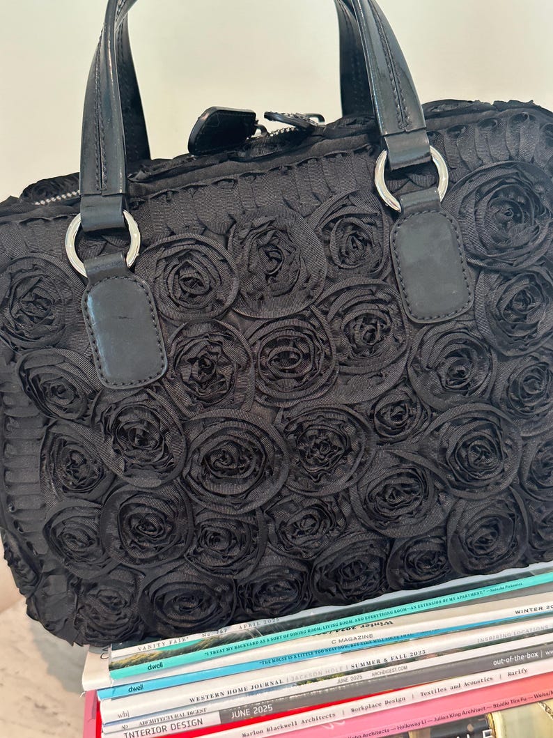 May include: Black handbag featuring a rose pattern. The bag has two handles with silver rings and a zip closure. It rests on a stack of magazines. The magazines have various titles.