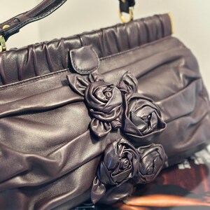May include: A dark purple handbag with a top handle and gold-colored hardware. The bag features a decorative arrangement of fabric roses on the front. The bag is made of a soft, pleated material.