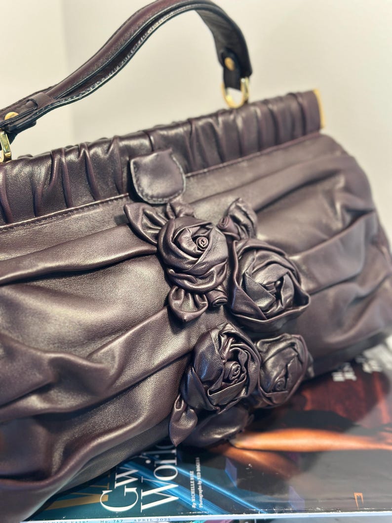 May include: A dark purple handbag with a top handle and gold-colored hardware. The bag features a decorative arrangement of fabric roses on the front. The bag is made of a soft, pleated material.