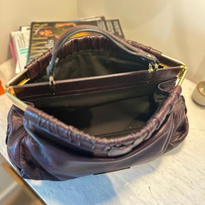 May include: A deep purple leather handbag with a gold-toned frame and handle. The bag is open, revealing a black interior. The leather has a gathered, ruched design. The bag sits on a white marble surface.