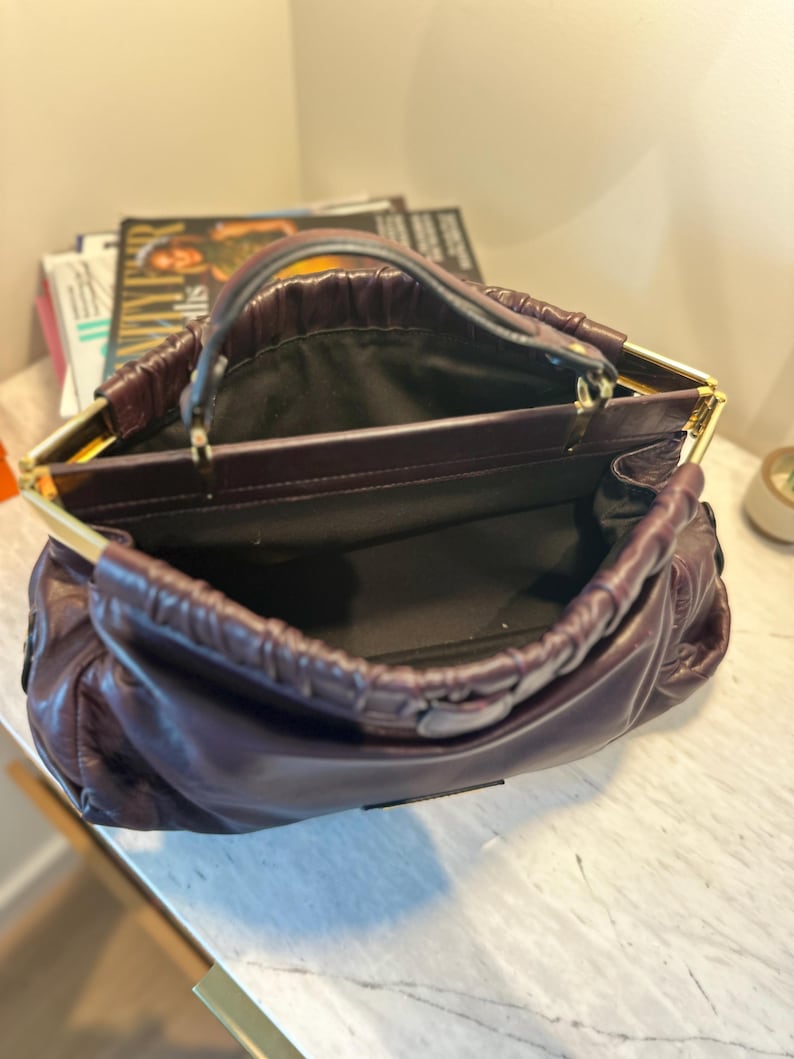 May include: A deep purple leather handbag with a gold-toned frame and handle. The bag is open, revealing a black interior. The leather has a gathered, ruched design. The bag sits on a white marble surface.
