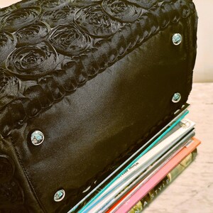 May include: Black handbag with a rose-patterned top and silver-coloured studs. The bag is resting on a stack of colourful magazines. The bag's design features a textured, floral pattern.