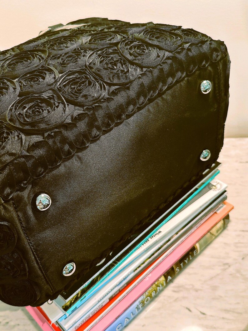 May include: Black handbag with a rose-patterned top and silver-coloured studs. The bag is resting on a stack of colourful magazines. The bag's design features a textured, floral pattern.