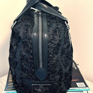 May include: Black handbag with a rose design, silver zip, and leather handles. The rectangular bag has a Valentino Garavani label. It sits on a stack of magazines.