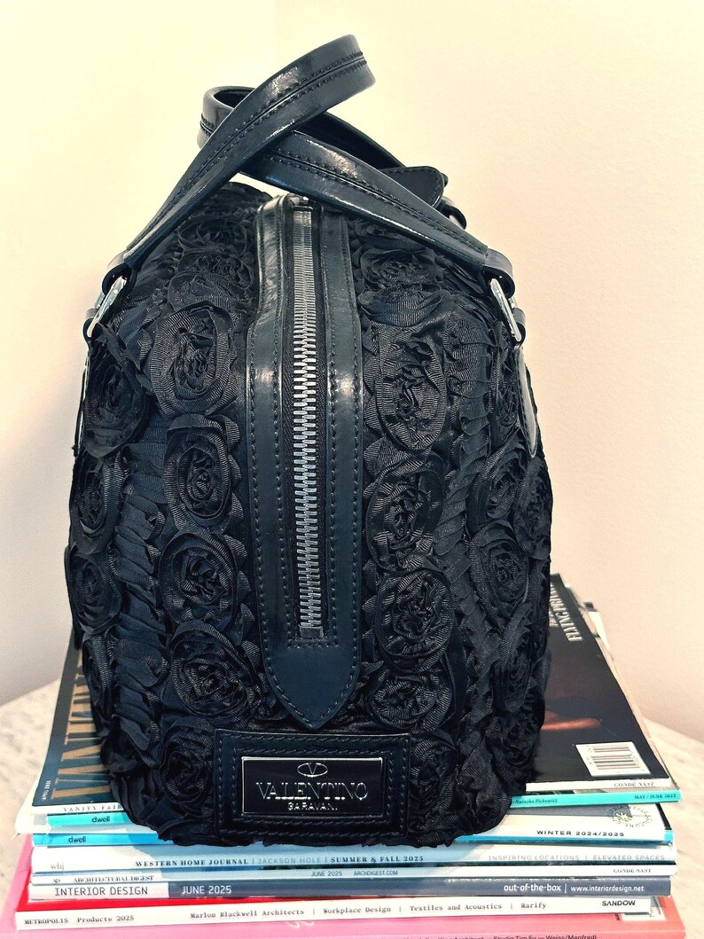 May include: Black handbag with a rose design, silver zip, and leather handles. The rectangular bag has a Valentino Garavani label. It sits on a stack of magazines.