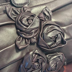 May include: Close-up of a dark brown leather handbag with a decorative floral design. The bag features three-dimensional leather roses and a pleated texture. The roses are intricately crafted, adding a touch of elegance to the accessory.
