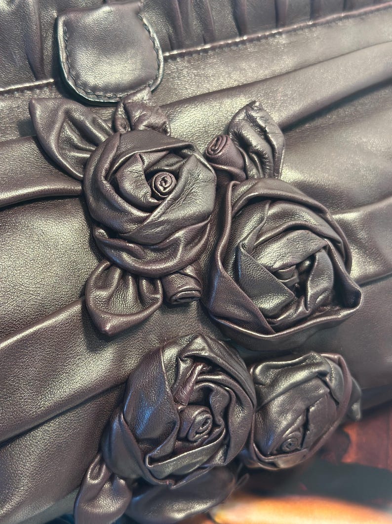 May include: Close-up of a dark brown leather handbag with a decorative floral design. The bag features three-dimensional leather roses and a pleated texture. The roses are intricately crafted, adding a touch of elegance to the accessory.