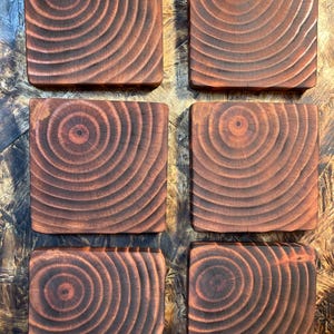 May include: Six square wooden coasters with a dark brown, burnt wood appearance. Each coaster features a unique circular grain pattern, showcasing the natural wood texture. The coasters are arranged on a textured surface.
