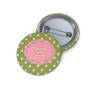 May include: A round button with a green background and pink polka dots. The button features a pink oval with the words "Matcha and joints" in orange script, framed by a decorative pink border. A second button is partially visible.