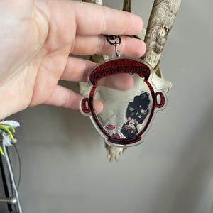 May include: Clear acrylic keychain featuring a red and black design of a character inside a decorative vessel. The keychain has a black metal ring for attachment. The design is visible through the clear acrylic.