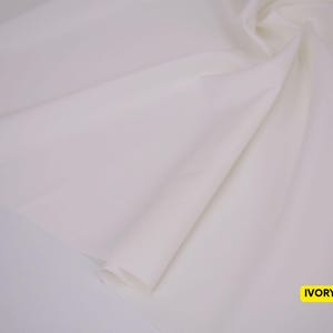 May include: A length of smooth, solid ivory-colored fabric draped in soft, flowing folds. The material has a matte finish and a consistent, light off-white tone throughout. A small yellow rectangular label in the corner displays the text IVORY.