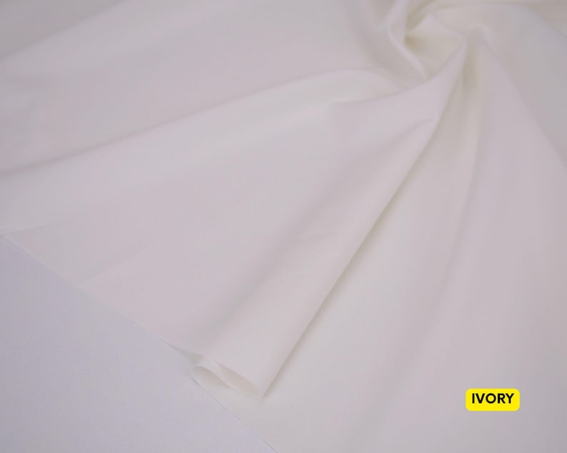May include: A length of smooth, solid ivory-colored fabric draped in soft, flowing folds. The material has a matte finish and a consistent, light off-white tone throughout. A small yellow rectangular label in the corner displays the text IVORY.
