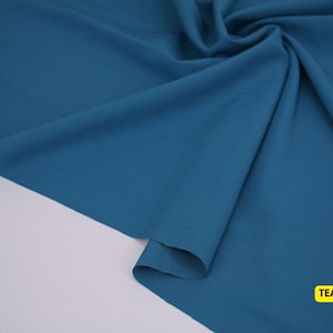 May include: A solid teal fabric with a smooth, matte texture, draped in soft folds to create natural shadows and highlights. The material is opaque and consistent in color throughout, with a small yellow rectangular label reading TEAL.