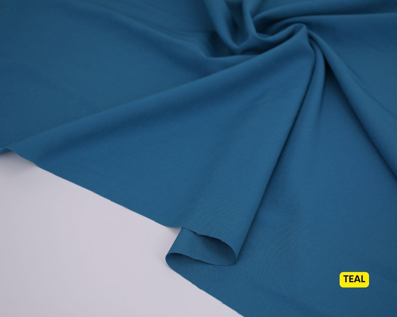 May include: A solid teal fabric with a smooth, matte texture, draped in soft folds to create natural shadows and highlights. The material is opaque and consistent in color throughout, with a small yellow rectangular label reading TEAL.