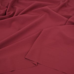 May include: A solid dark red fabric with a smooth, matte texture. The material is draped in soft, flowing folds, creating deep shadows and highlights across the surface. The fabric appears opaque and consistent in its deep burgundy red color.